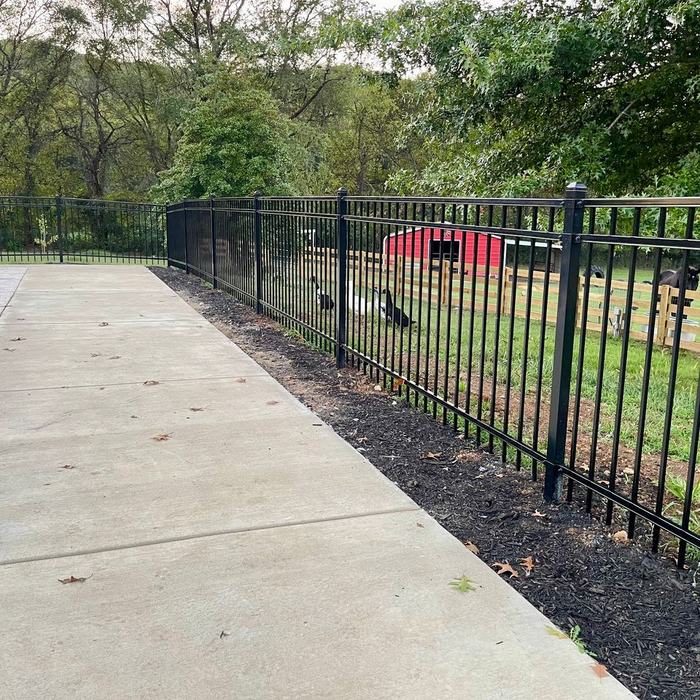 Black ornamental fence running beside a concrete sidewalk toward a barn