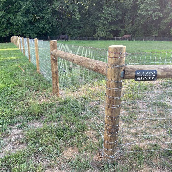Woven wire livestock fence on wood posts with a Meadows Farm and Fence sign