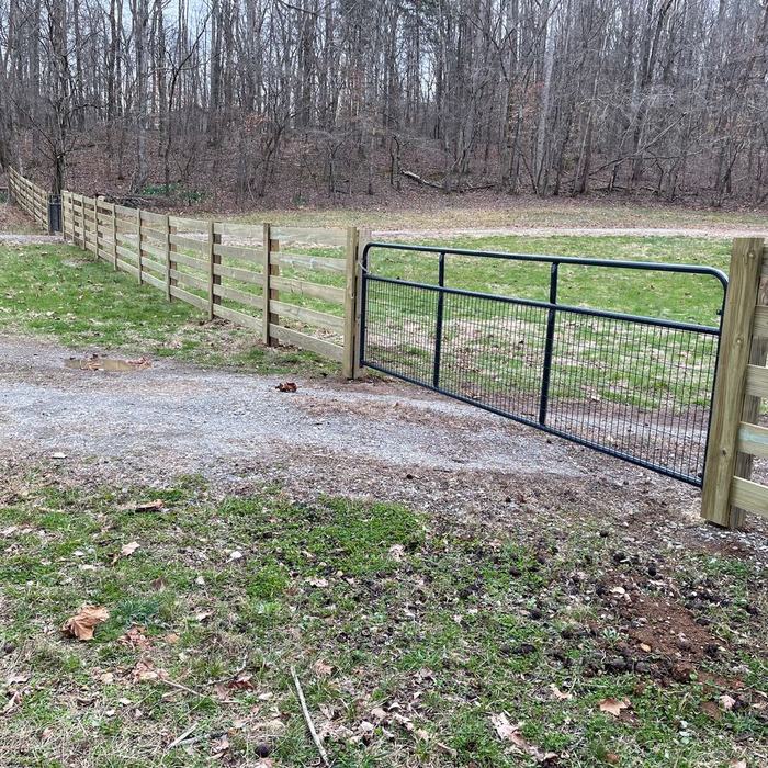 Black metal farm gate hung between rail fence ends at a gravel drive entrance